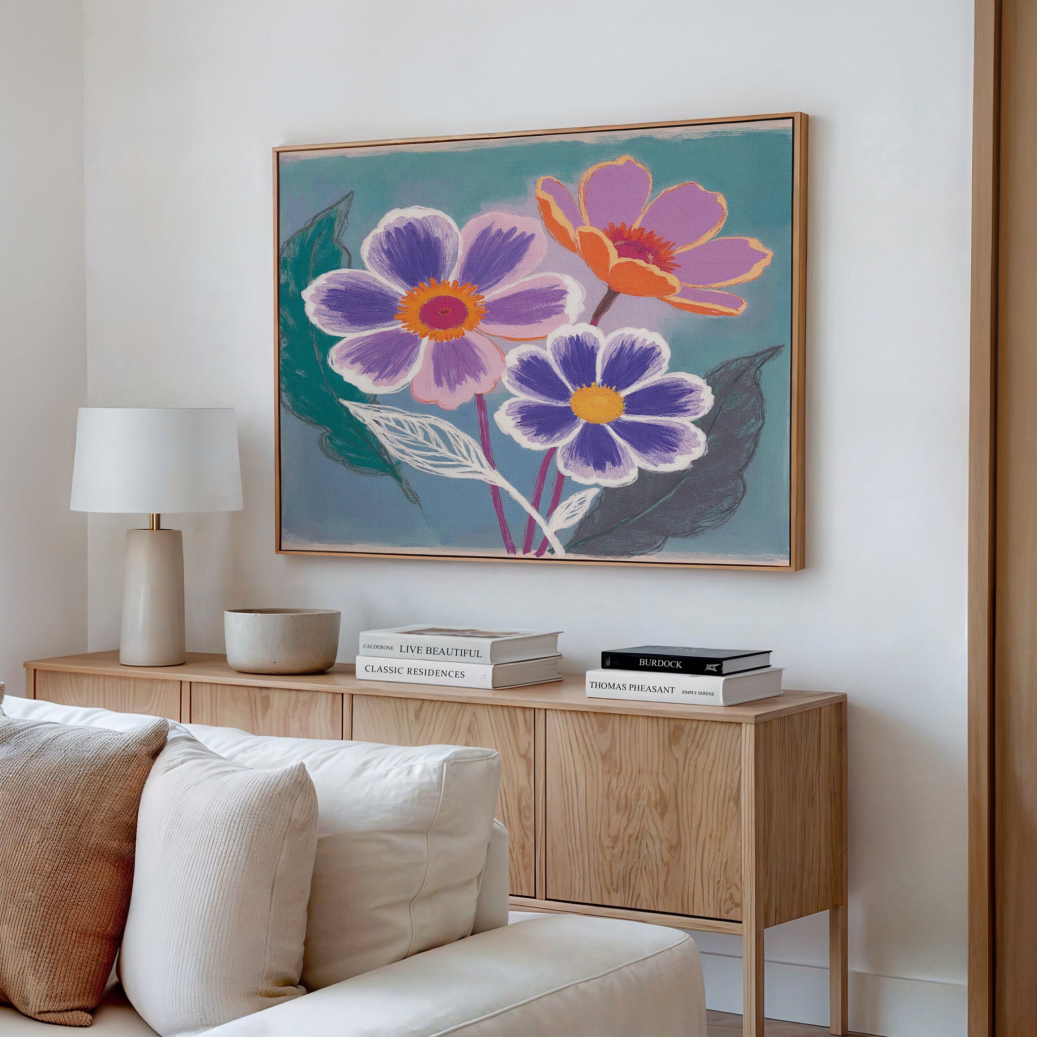 A modern living room showcases a wooden sideboard with stacked books, a white lamp, a bowl, and the Vibrant Petals Canvas Art - Horizontal featuring colorful flowers above it. Neutral-toned cushions appear in the foreground.