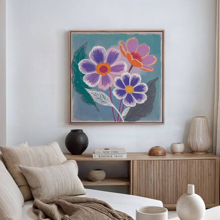 A cozy living room with a neutral sofa, beige and cream cushions, vases, stacked books, and a wooden sideboard. Above, the Vibrant Petals Canvas Art - Square adds Nordic Park-inspired color with its three large flowers.