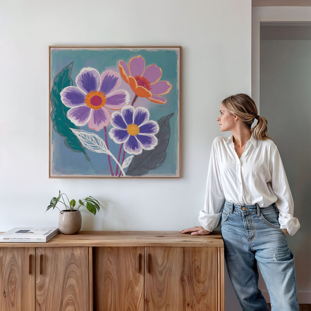 A woman in a white shirt and jeans leans against a wooden cabinet, gazing at the Vibrant Petals Canvas Art - Square above her. A small potted plant and books rest on the cabinet, creating a cozy Nordic Park-inspired scene.