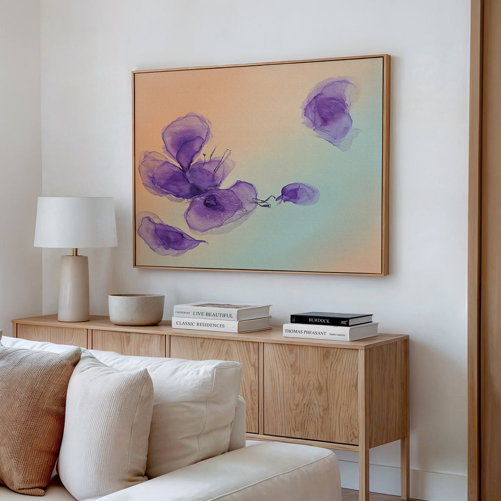 A modern living room with a light wood sideboard, stacked books, a ceramic bowl, a white lamp, and the Violet Dreams Canvas Art - Horizontal featuring abstract purple flowers on peach above a pale sofa.