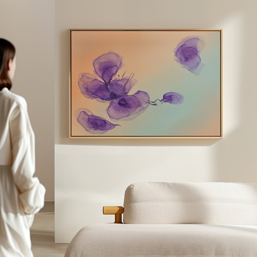 A person in a white outfit stands in a bright, minimal room, admiring the Violet Dreams Canvas Art - Horizontal featuring abstract purple flowers on a pastel background above a cream sofa.