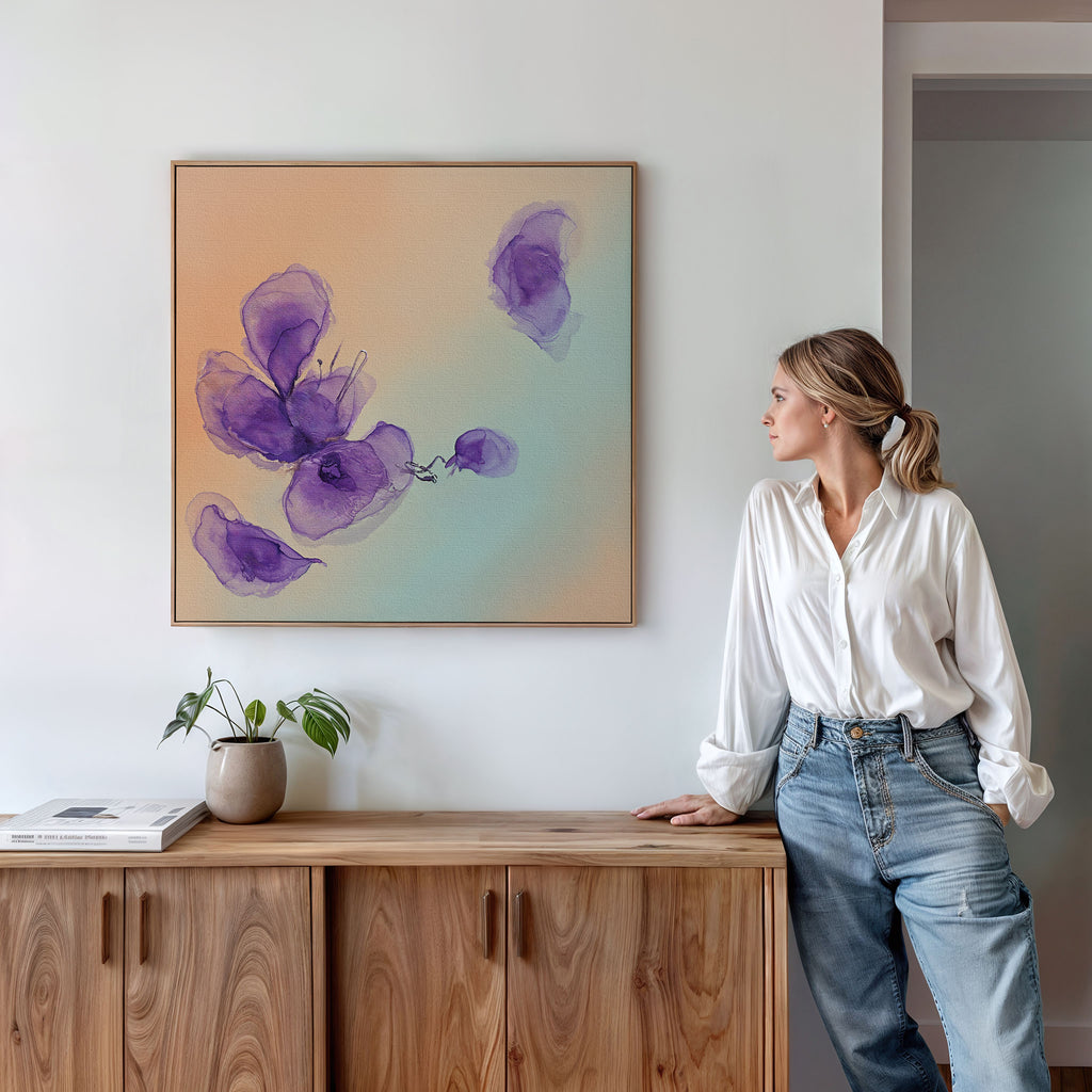 A woman in a white blouse and jeans stands by a wooden sideboard, admiring the Violet Dreams Canvas Art - Square, featuring abstract purple flowers on a light wall. Botanical accents, books, and a potted plant complete the Nordic Park-inspired scene.