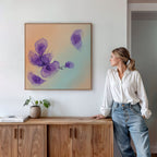 A woman in a white blouse and jeans stands by a wooden sideboard, admiring the Violet Dreams Canvas Art - Square, featuring abstract purple flowers on a light wall. Botanical accents, books, and a potted plant complete the Nordic Park-inspired scene.