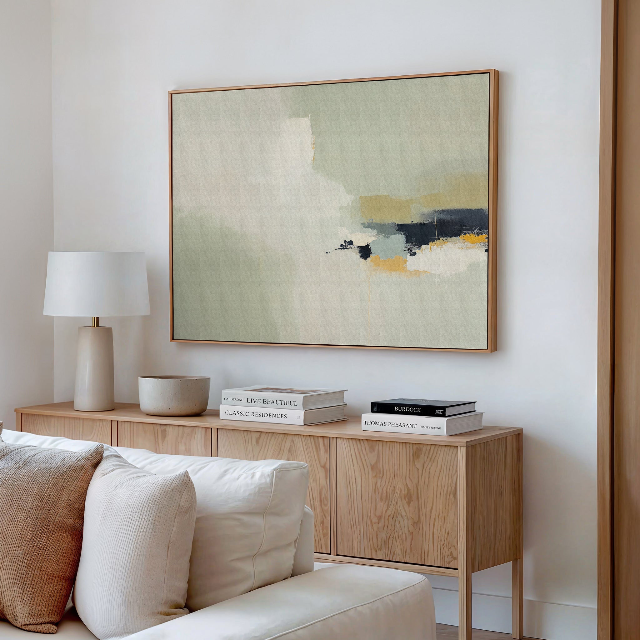 A modern living room features a light wood sideboard with stacked books, a lamp, and a bowl, while the Weathered Composition Canvas Art - Horizontal in neutral tones hangs above a beige sofa with white and cream pillows for an elegant look.