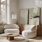 A modern living room featuring two rounded white chairs, a low wooden coffee table, and Weathered Composition - Set Of 2 abstract prints leaning against the wall, adding elegance beside a tall lamp and a large window with natural light.