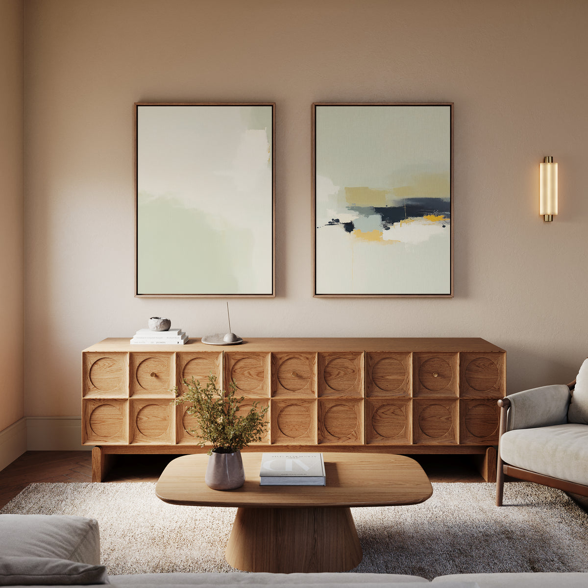 A modern living room with a light wood sideboard, round coffee table, beige armchair, and "Weathered Composition - Set Of 2" abstract prints on a light wall. A small plant, books, and wall sconce complete the elegant modern decor.
