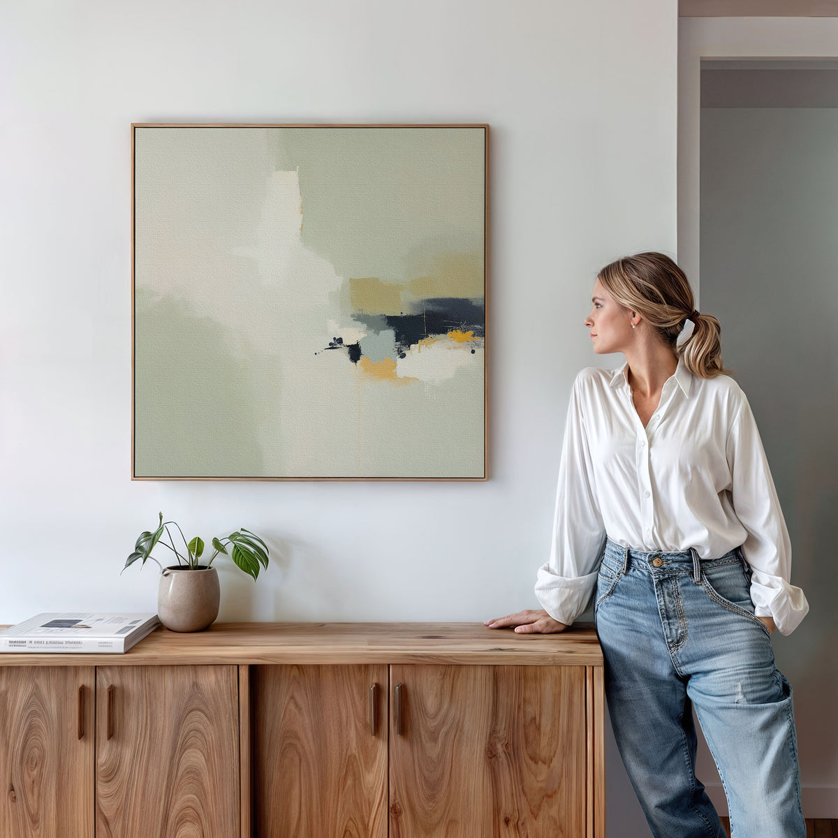 A woman in a white blouse and jeans leans against a wooden cabinet, admiring the Weathered Composition Canvas Art - Square above. A small plant and books on the cabinet add to the room’s modern, minimalist elegance.