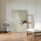 A minimalist living space features a white cushioned chair, a round black side table with candles and books, and the Weathered Composition Canvas Art - Vertical resting against a paneled white wall on a parquet wood floor.