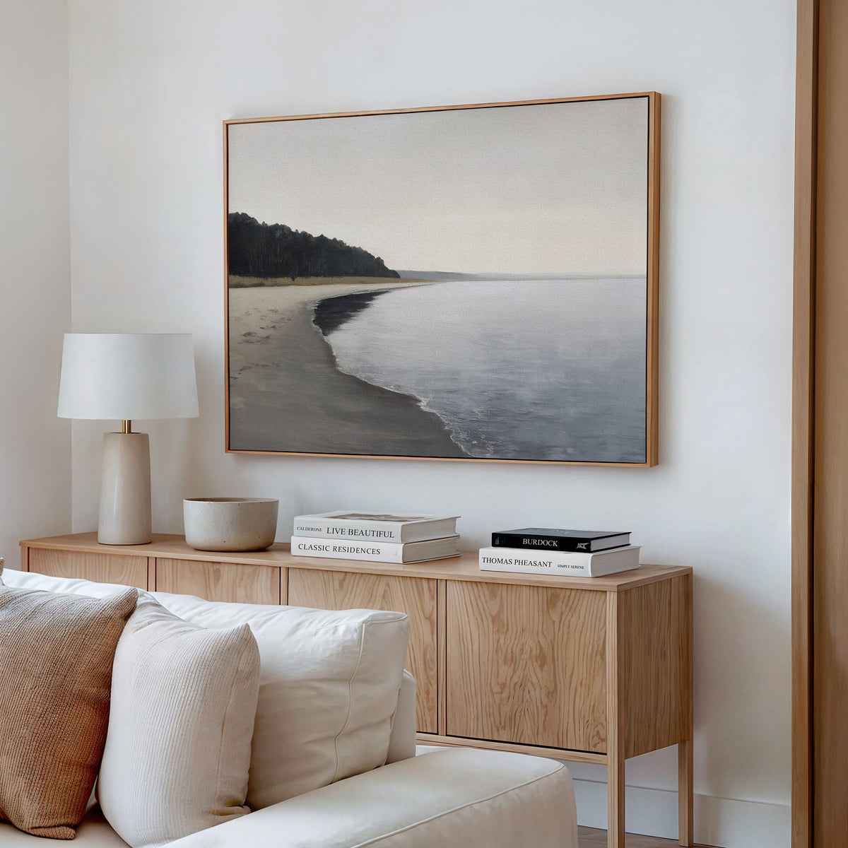 A minimalist living room with a wooden sideboard, neutral decor, books, a white lamp, and the Weathered Shore Canvas Art - Horizontal displayed above the sideboard on a white wall.