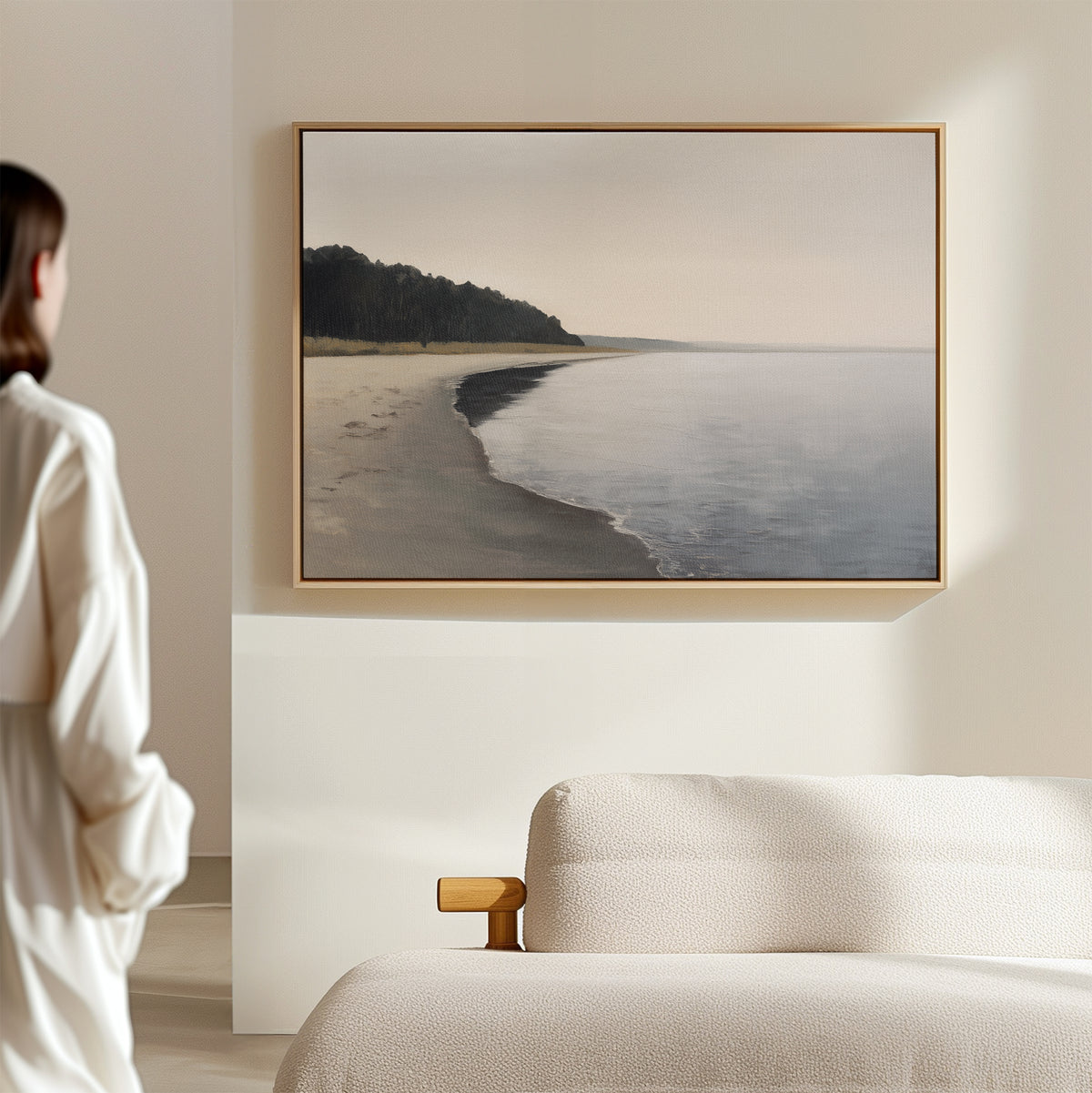 A person stands in a softly lit, modern room, admiring the Weathered Shore Canvas Art - Horizontal, which depicts a calm shoreline with distant trees and is elegantly displayed above a light-colored sofa.