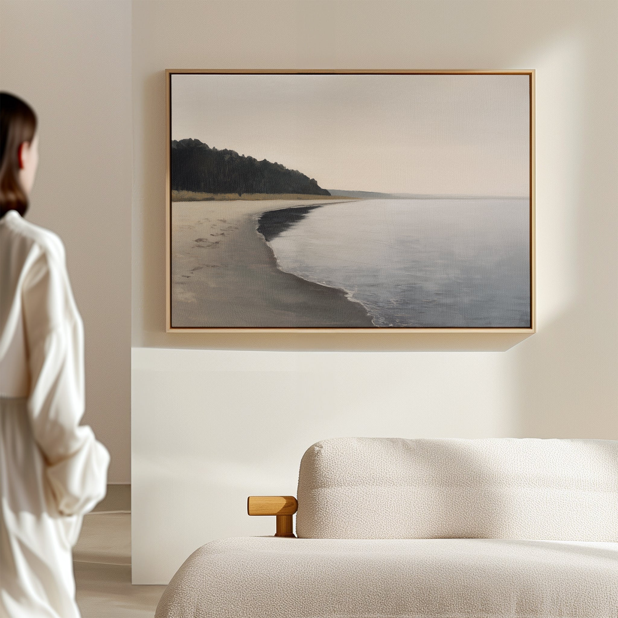A person stands in a softly lit, modern room, admiring the Weathered Shore Canvas Art - Horizontal, which depicts a calm shoreline with distant trees and is elegantly displayed above a light-colored sofa.