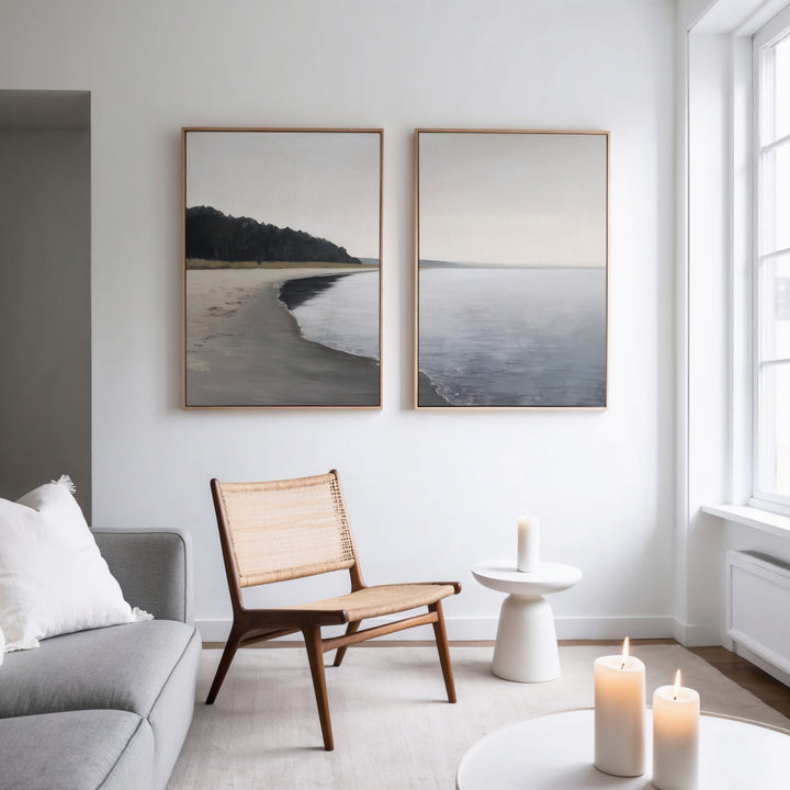 A minimal, modern living room with a woven chair, small white table, lit candles, grey sofa, and the Weathered Shore - Set of 2 wall art on a white wall near a large window.
