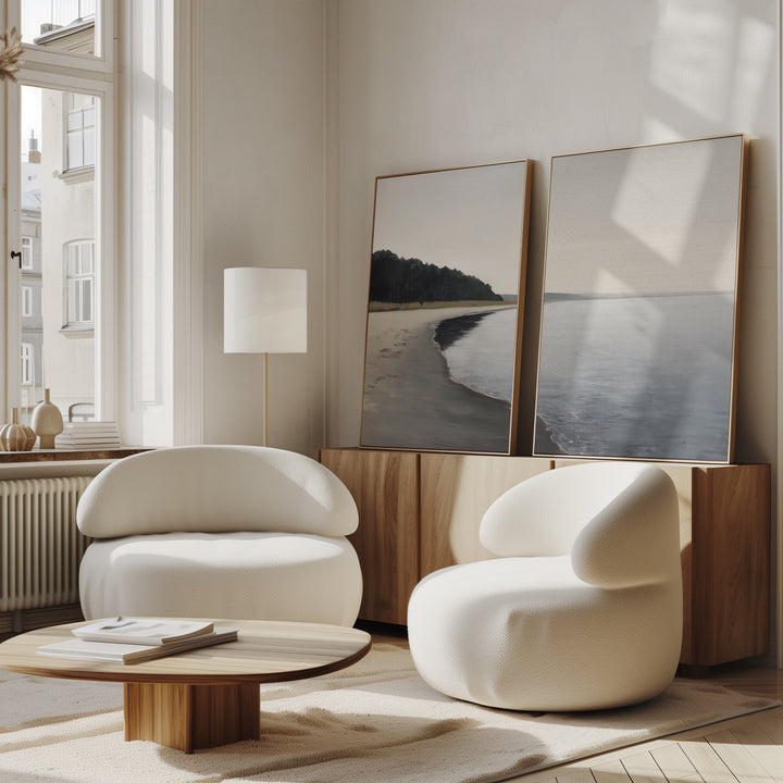 Two modern white armchairs and a low wooden table sit in a bright living room. Large windows, a floor lamp, and the Weathered Shore - Set of 2 wall art leaning on a wooden cabinet create a calm, minimalist feel.