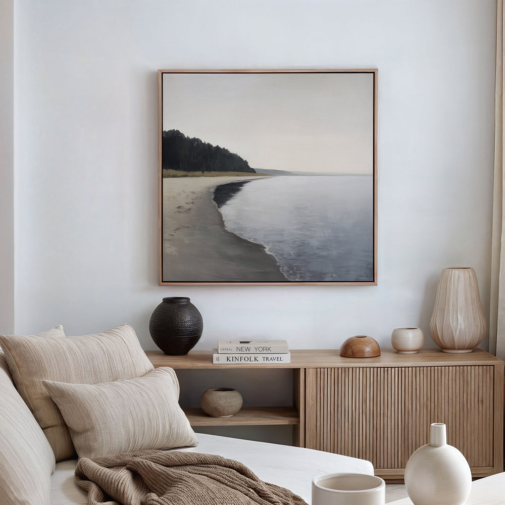 A serene, minimalist living room features neutral-toned furniture and decor. Above a wooden sideboard with books and vases hangs the Weathered Shore Canvas Art - Square, showcasing a calm shoreline for a relaxing coastal vibe.