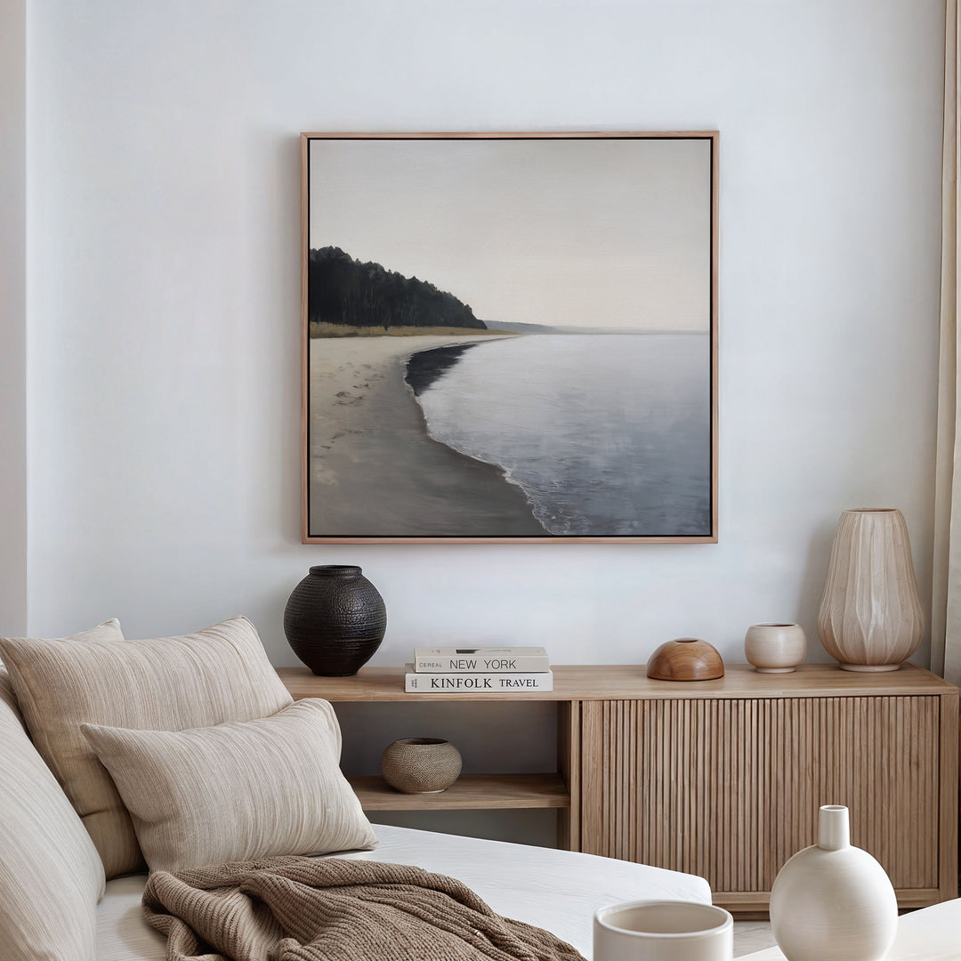 A serene, minimalist living room features neutral-toned furniture and decor. Above a wooden sideboard with books and vases hangs the Weathered Shore Canvas Art - Square, showcasing a calm shoreline for a relaxing coastal vibe.