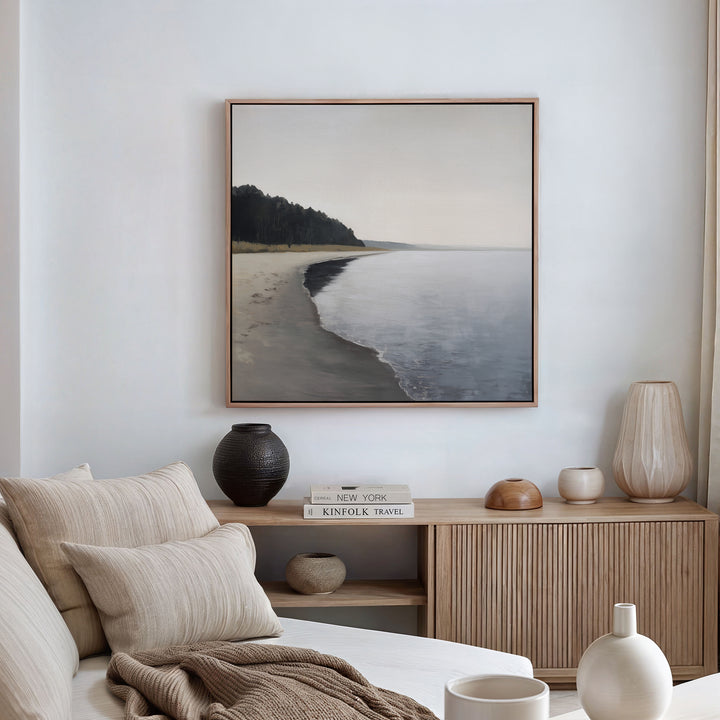 A serene, minimalist living room features neutral-toned furniture and decor. Above a wooden sideboard with books and vases hangs the Weathered Shore Canvas Art - Square, showcasing a calm shoreline for a relaxing coastal vibe.