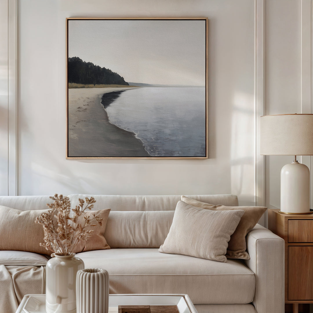 A neutral-toned living room features a beige sofa, cream pillows, a side table with a lamp, and dried flowers in a vase. Above the sofa hangs the Weathered Shore Canvas Art - Square, bringing serene coastal vibes to the space.