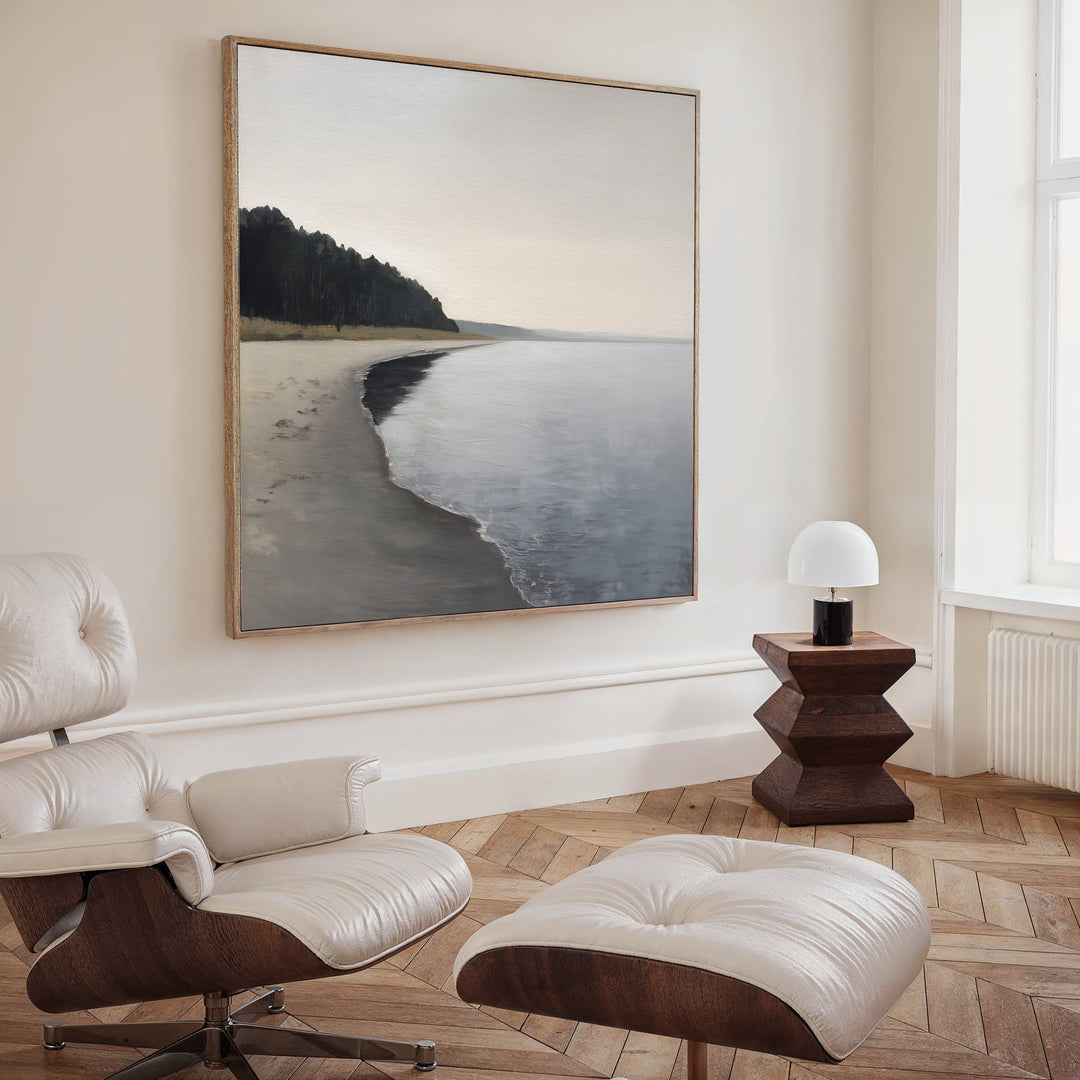 A modern minimalist living room features the Weathered Shore Canvas Art - Square, a white leather lounge chair with ottoman, a wooden geometric side table, and a small white lamp by a tall window.
