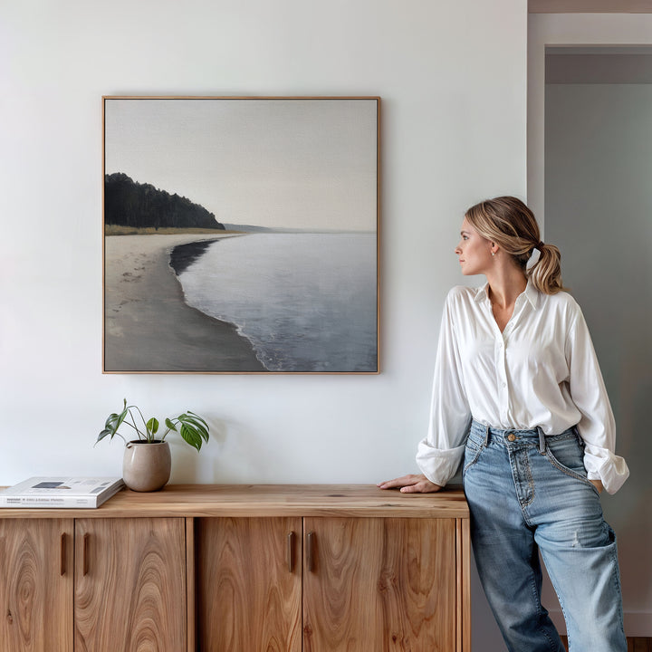 A woman in a white blouse and jeans stands by a wooden sideboard, admiring the Weathered Shore Canvas Art - Square on a white wall. A potted plant and books on the sideboard enhance the relaxed, coastal vibe.