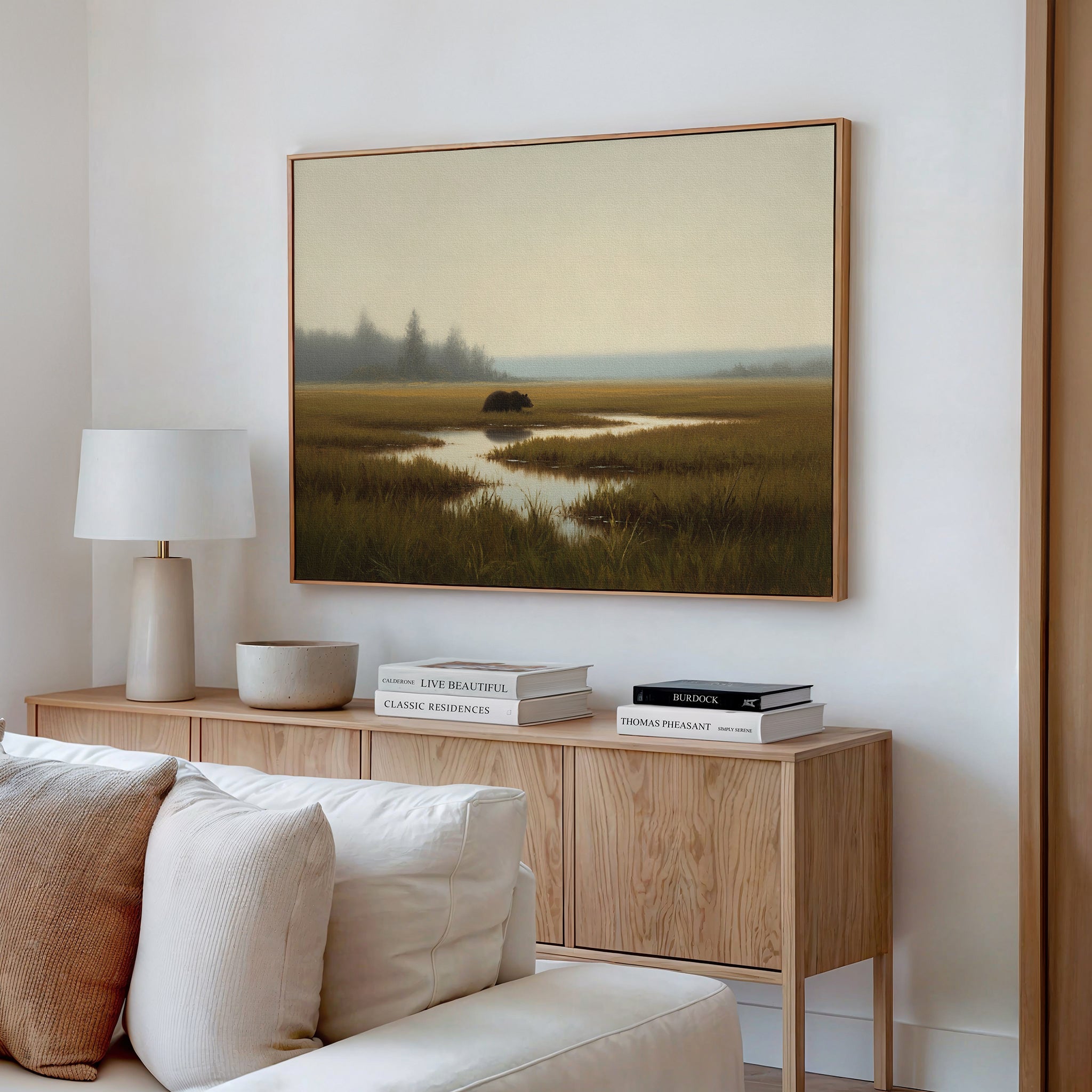 A modern living room with a light wood console, white lamp, neutral decor, and a white sofa. Above the console hangs the Wetland Bear Canvas Art - Horizontal featuring a tranquil misty marsh with distant trees and calm water.