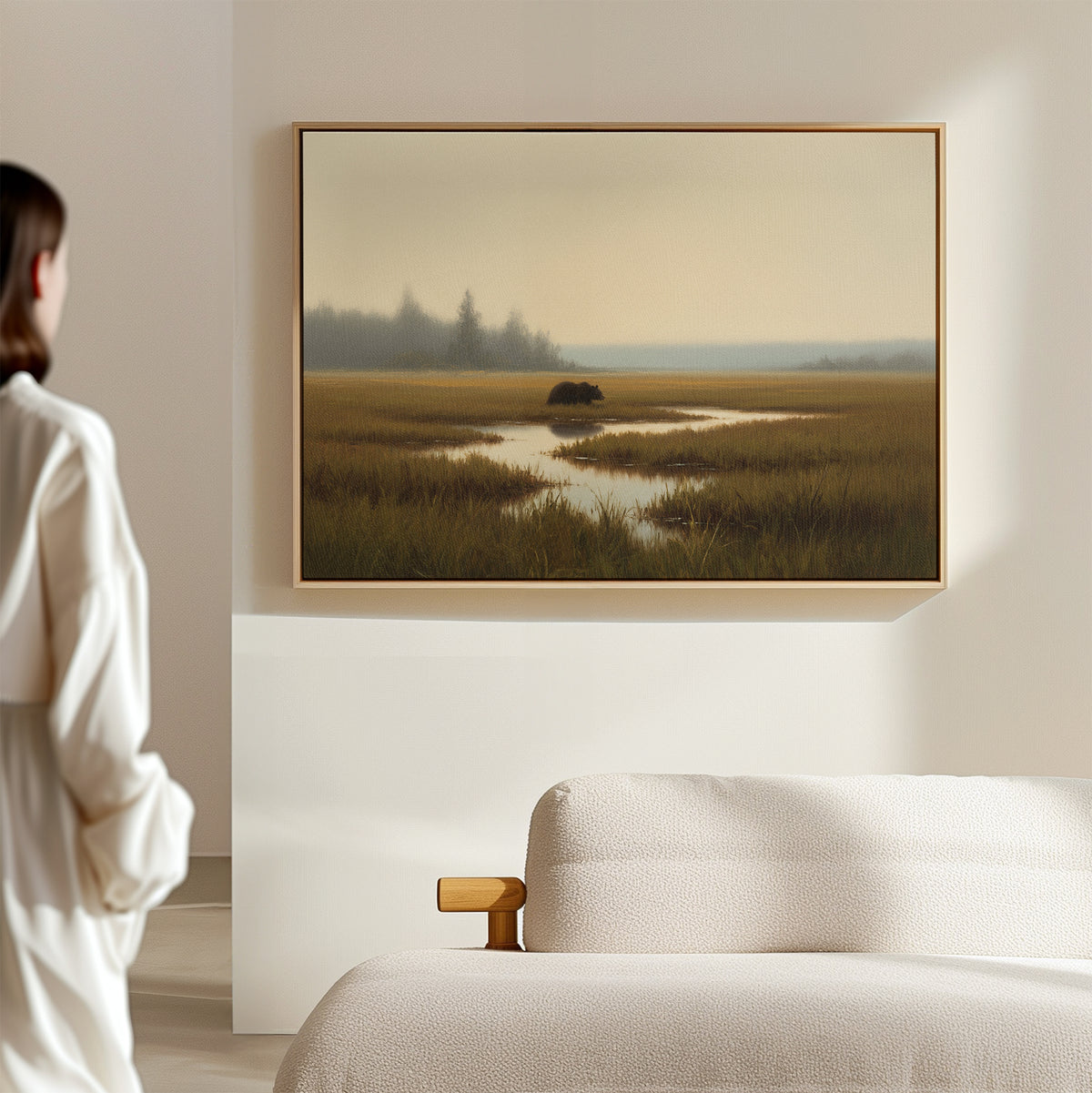 A person stands in a minimalistic, sunlit room, facing the Wetland Bear Canvas Art - Horizontal—a serene marshland scene with tall grass and water—displayed above a plush white sofa.