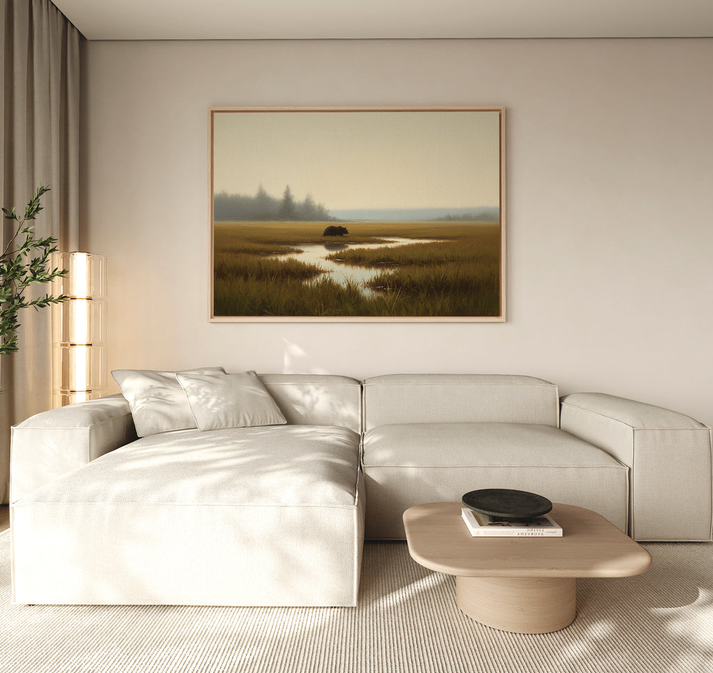 A modern living room featuring a light beige sectional sofa, wooden coffee table with a black bowl, tall plant, and the Wetland Bear Canvas Art - Horizontal depicting serene river and fields. Neutral tones and natural light create a tranquil atmosphere.
