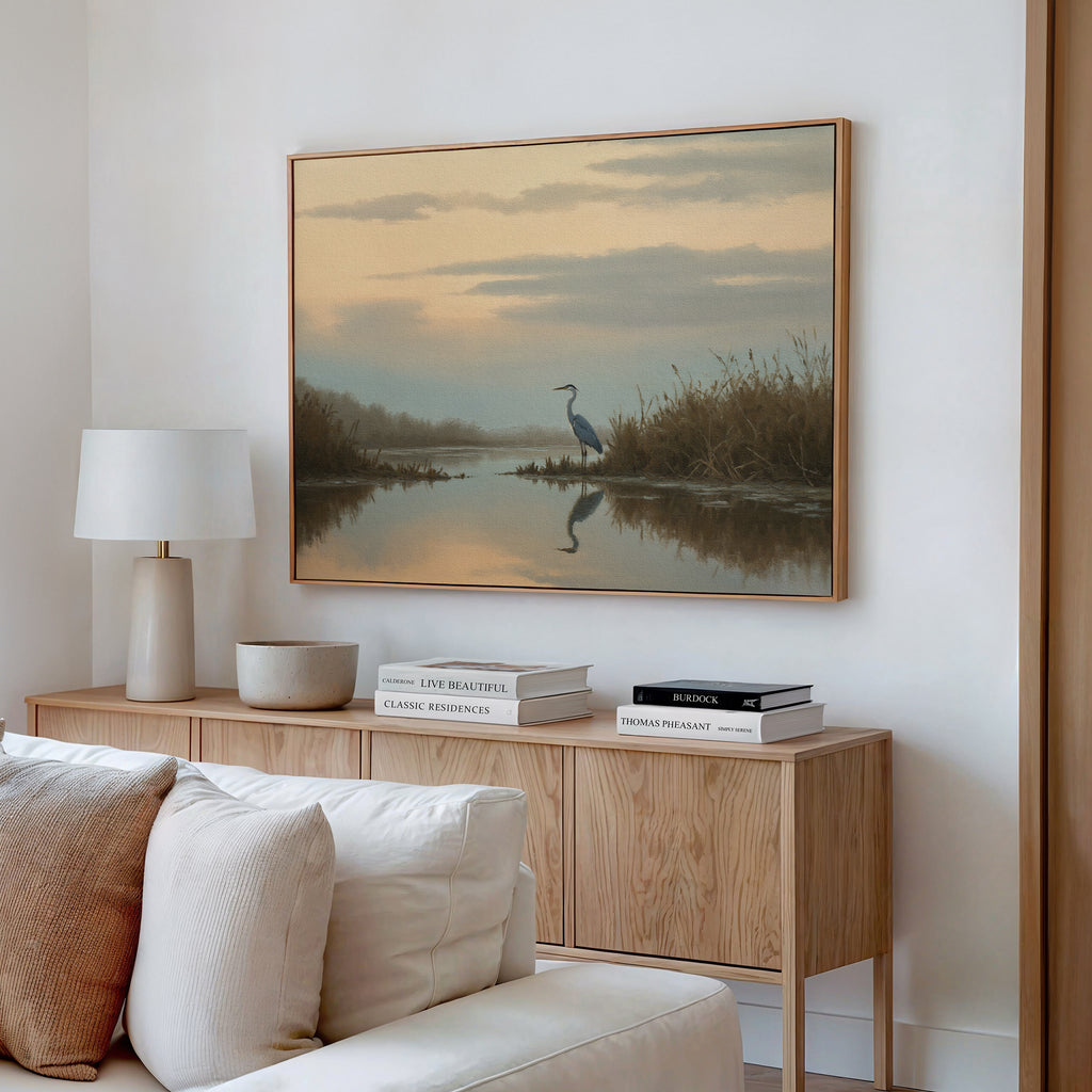 A serene living room features a light wood console, white lamp, books, and bowl. Above, the Wetland Heron Canvas Art - Horizontal depicting a misty marsh hangs on a white wall. A cream sofa sits in the foreground.