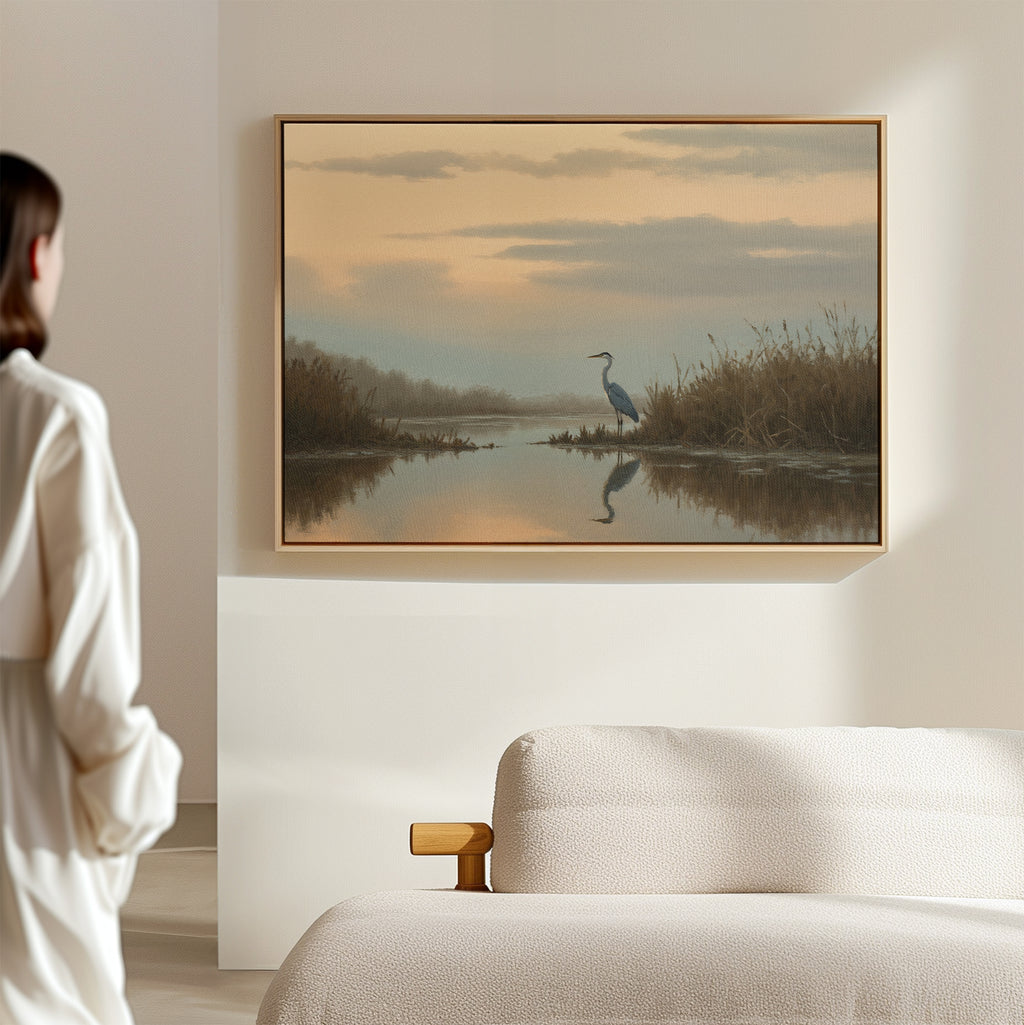 A person stands in a softly lit room, admiring the Wetland Heron Canvas Art - Horizontal, which depicts a heron by a sunset-lit waters edge with reeds and clouds reflected in the calm water, hung above a beige couch.