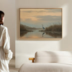 A person stands in a softly lit room, admiring the Wetland Heron Canvas Art - Horizontal, which depicts a heron by a sunset-lit waters edge with reeds and clouds reflected in the calm water, hung above a beige couch.