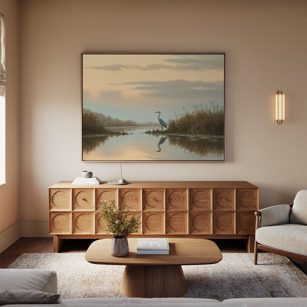 A cozy living room features a wooden sideboard and coffee table, a beige sofa, a wall sconce, and the Wetland Heron Canvas Art - Horizontal print depicting a serene heron at sunset hanging above the sideboard.