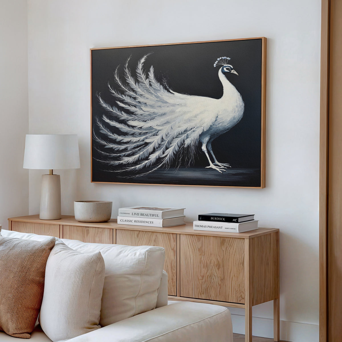 A modern living room with a light wood sideboard, white lamp, stacked books, and elegant wall art—a White Peacock Display Canvas Art - Horizontal—hanging above.