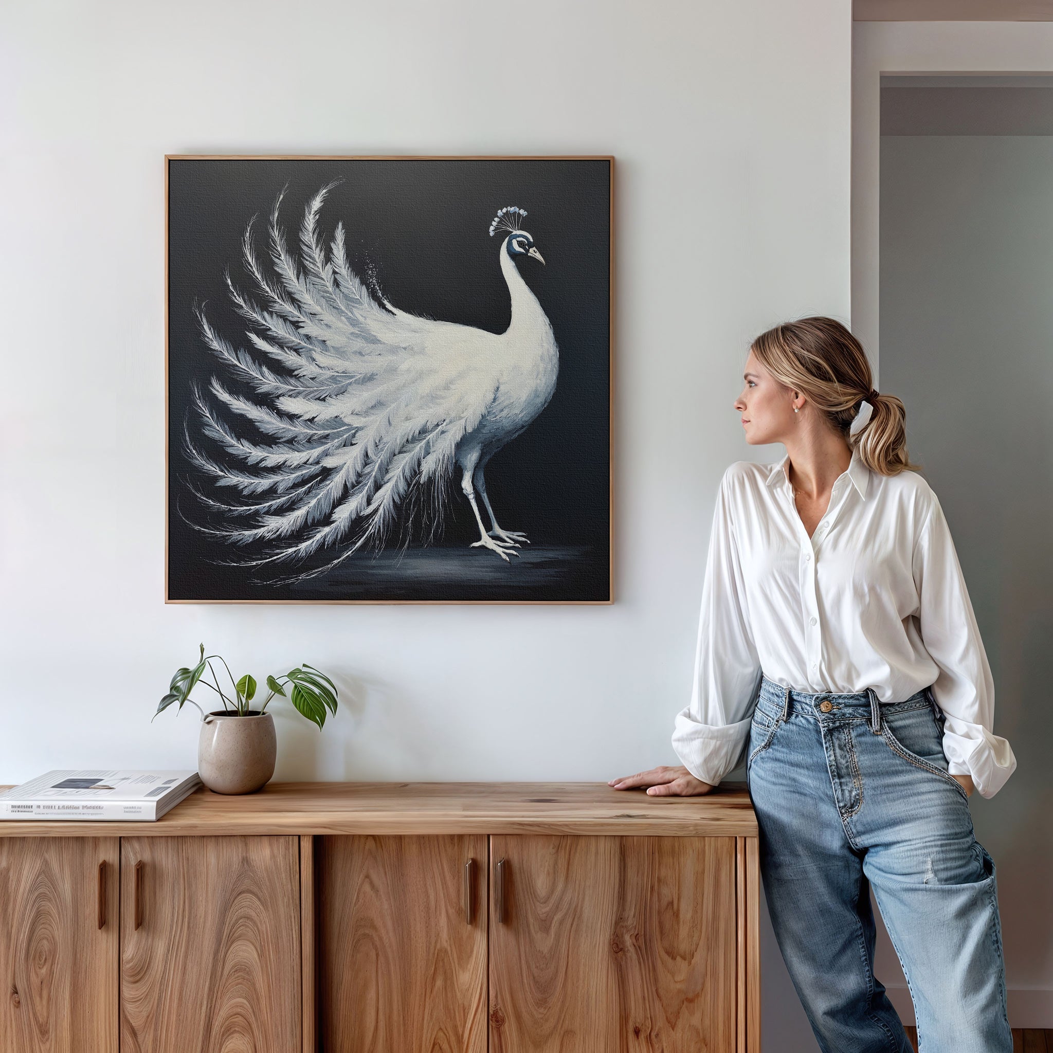 A woman in a white shirt and jeans leans against a wooden cabinet, gazing at the White Peacock Display Canvas Art - Square that hangs on the white wall. A potted plant and books add decor to the cabinet.