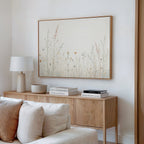A minimalist living room features a light wood sideboard, white table lamp, books, and decor. Above, the Wildflower Dreams Canvas Art - Horizontal brings botanical flair to the white wall. A white couch with pillows sits invitingly in front.
