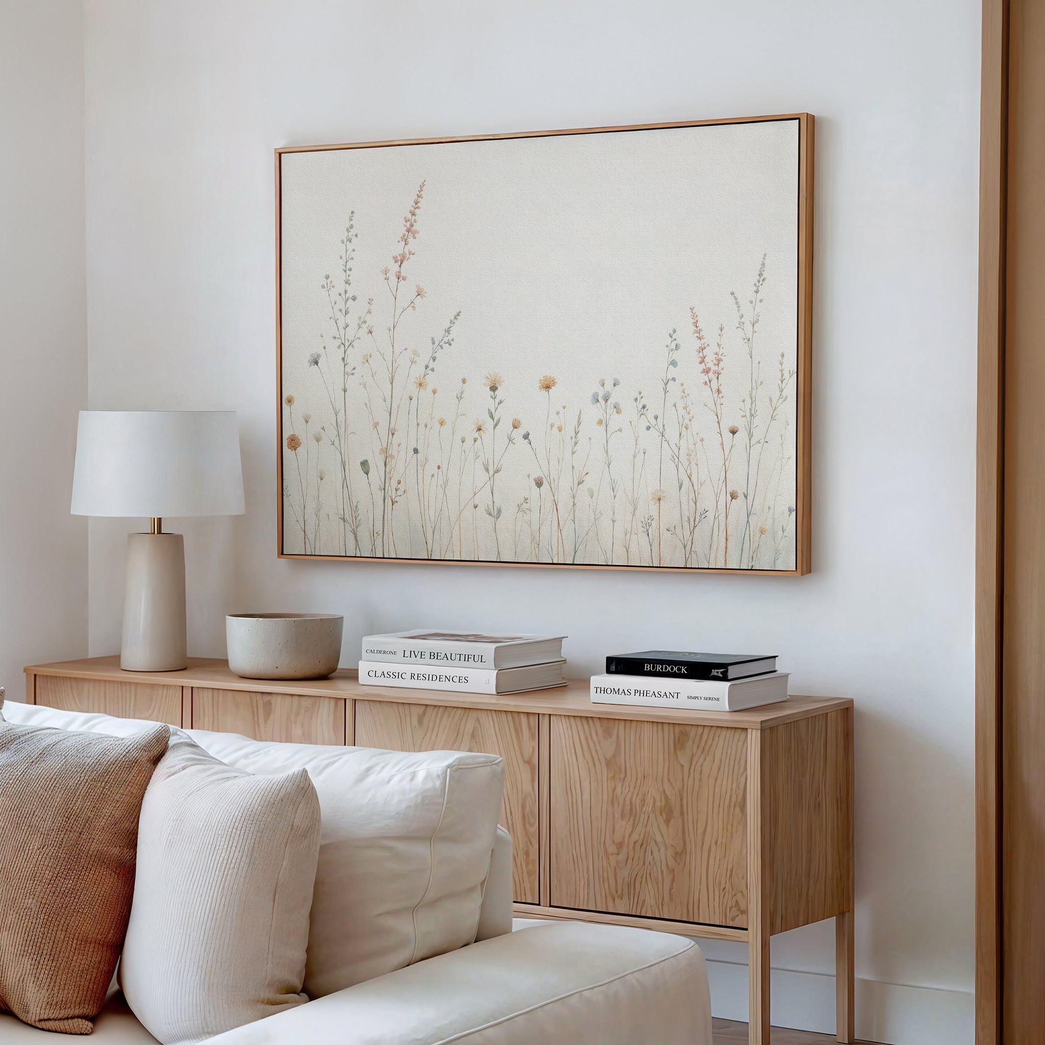 A minimalist living room features a light wood sideboard, white table lamp, books, and decor. Above, the Wildflower Dreams Canvas Art - Horizontal brings botanical flair to the white wall. A white couch with pillows sits invitingly in front.
