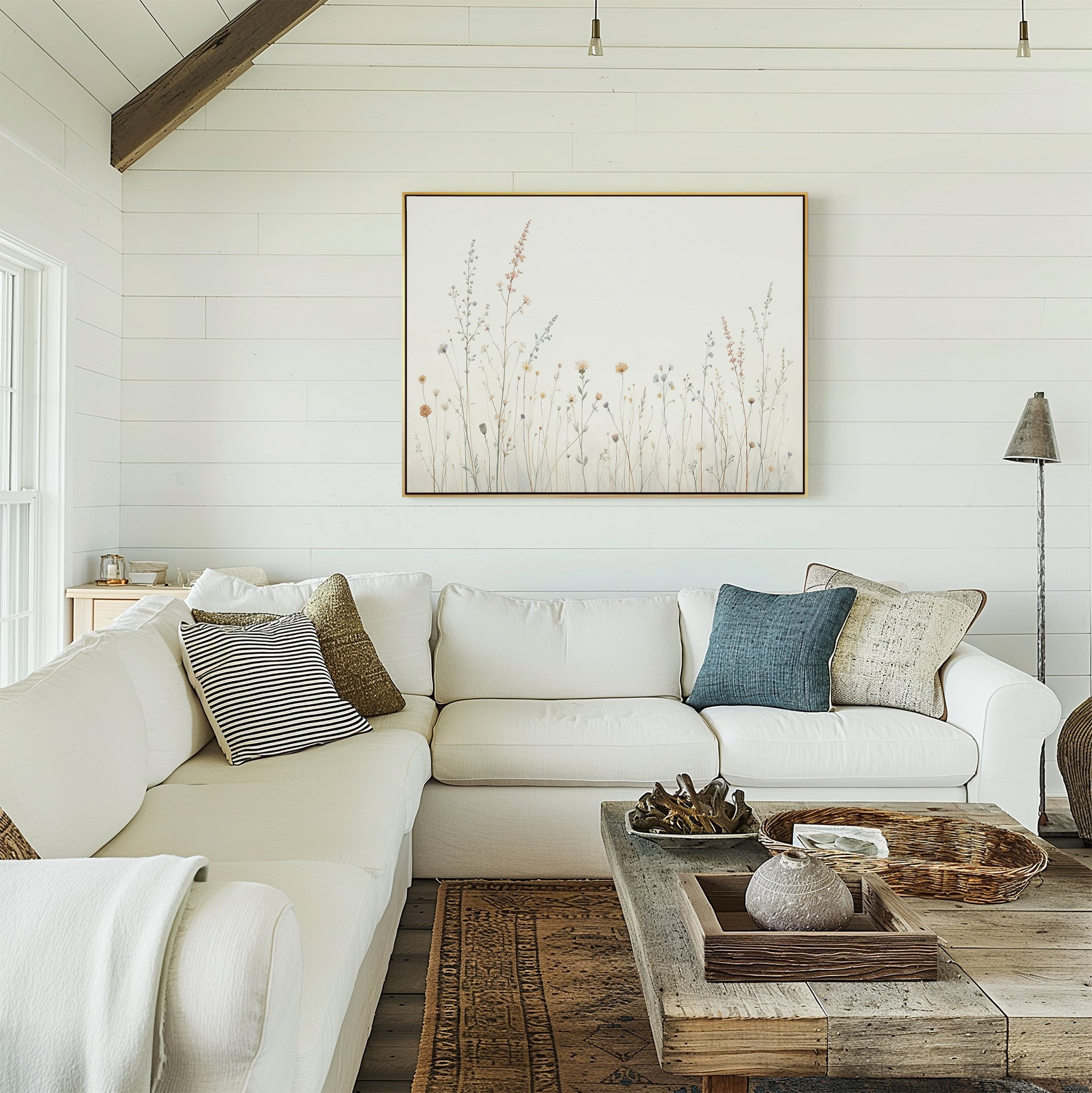 A cozy living room with a white sectional sofa, assorted pillows, a rustic wooden coffee table, and the Wildflower Dreams Canvas Art - Horizontal on a white shiplap wall. Natural light streams in from a window on the left.