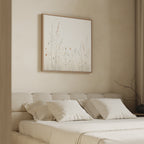A minimalist bedroom with a beige upholstered bed, white pillows and bedding, and the "Wildflower Dreams Canvas Art - Square" featuring delicate wildflowers above the headboard. A bare branch sits in a vase on the nightstand.