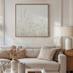 A neutral-toned living room features a beige sofa, matching cushions, a wooden side table with a cream lamp, and dried flowers in a vase. Above the sofa hangs Wildflower Dreams Canvas Art - Square, bringing nature into the minimalist space.