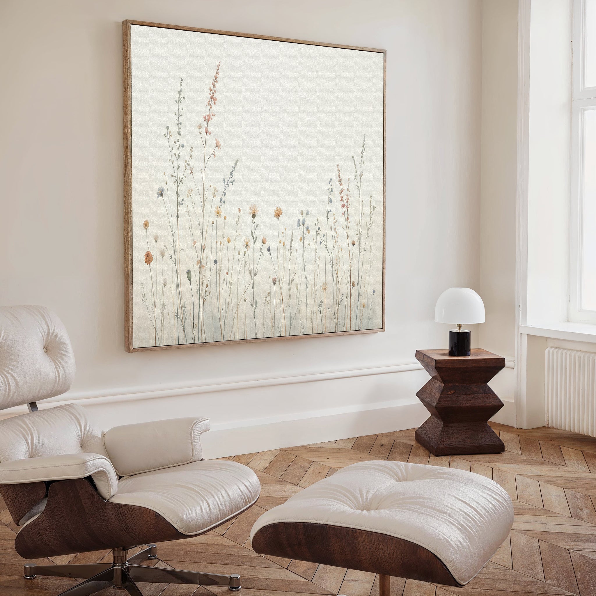 A modern, minimalist living room with a white lounge chair and ottoman, a geometric wooden side table with a small white lamp, and the Wildflower Dreams Canvas Art - Square as stunning botanical wall art on a cream wall.