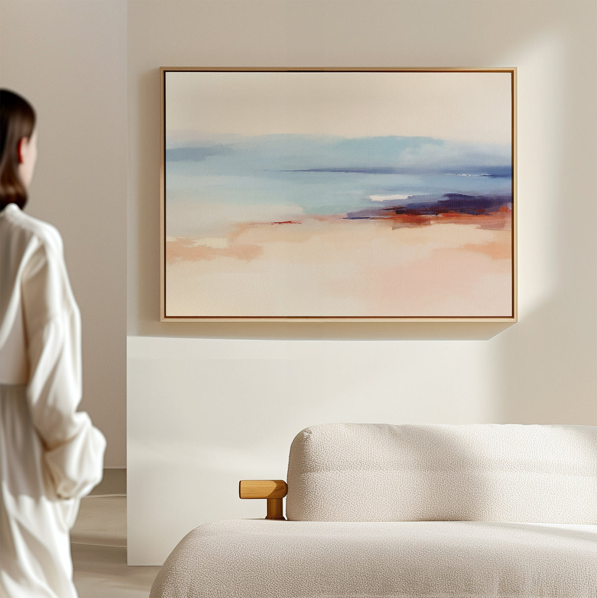 In a minimal, softly lit room, a person admires the Windswept Coastline Canvas Art - Horizontal, featuring pastel blue, peach, and beige tones above a light sofa—elevating the tranquil atmosphere with elegant coastal style.
