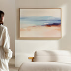 In a minimal, softly lit room, a person admires the Windswept Coastline Canvas Art - Horizontal, featuring pastel blue, peach, and beige tones above a light sofa—elevating the tranquil atmosphere with elegant coastal style.