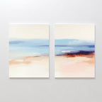 The "Windswept Coastline - Set Of 2" features abstract paintings in blue, beige, and peach tones, evoking a serene seascape. Hung side by side, these prints create elegant coastal wall art on a white wall.
