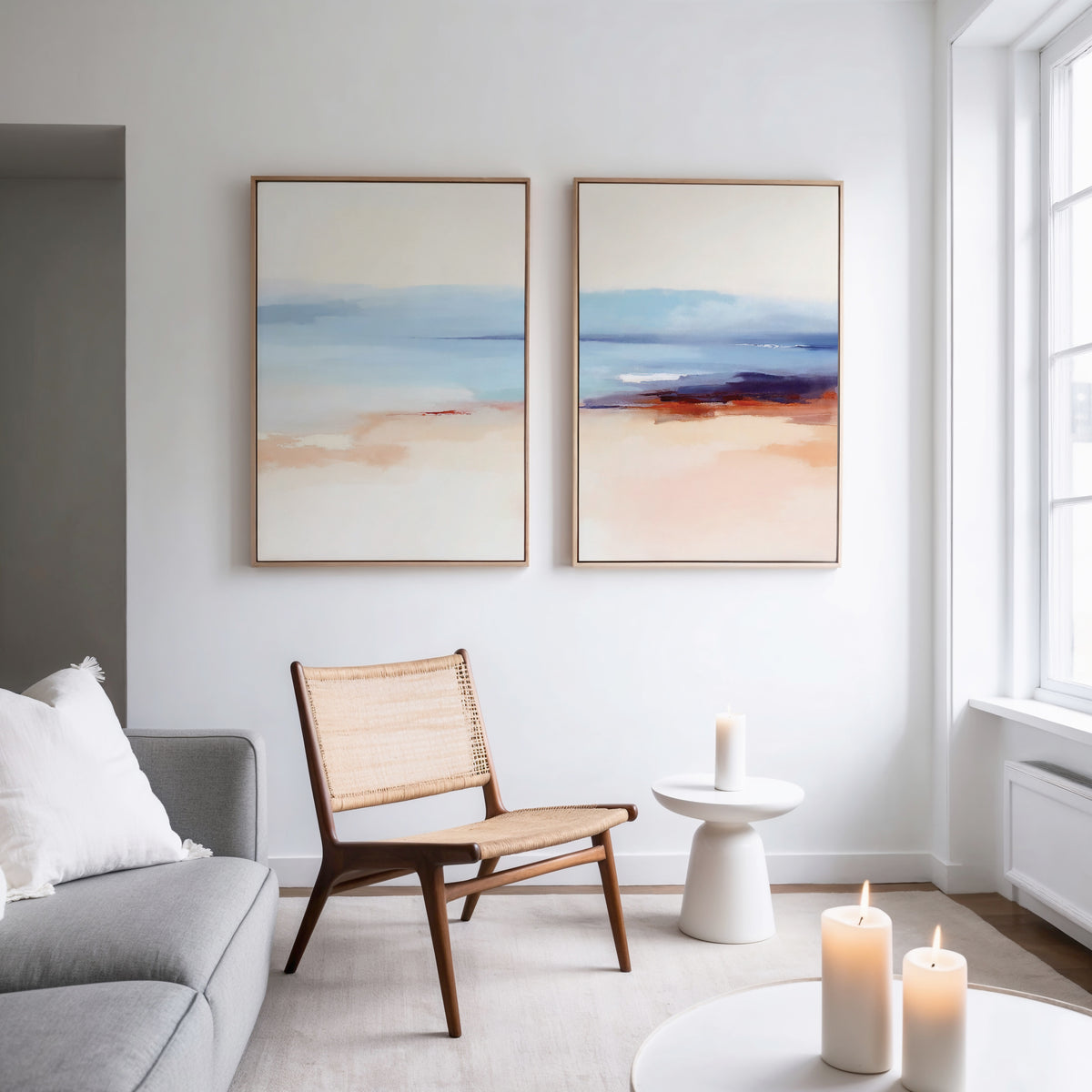 A modern, minimal living room with neutral tones features a woven chair, round side table with a candle, and the Windswept Coastline - Set Of 2 art prints, plus three candles on a white coffee table by a large window.