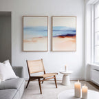 A modern, minimal living room with neutral tones features a woven chair, round side table with a candle, and the Windswept Coastline - Set Of 2 art prints, plus three candles on a white coffee table by a large window.