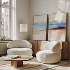 A modern living room features two rounded white armchairs, a low wooden coffee table, a floor lamp, and the Windswept Coastline - Set Of 2 wall art set atop a wooden sideboard against a white wall, brightened by large windows.