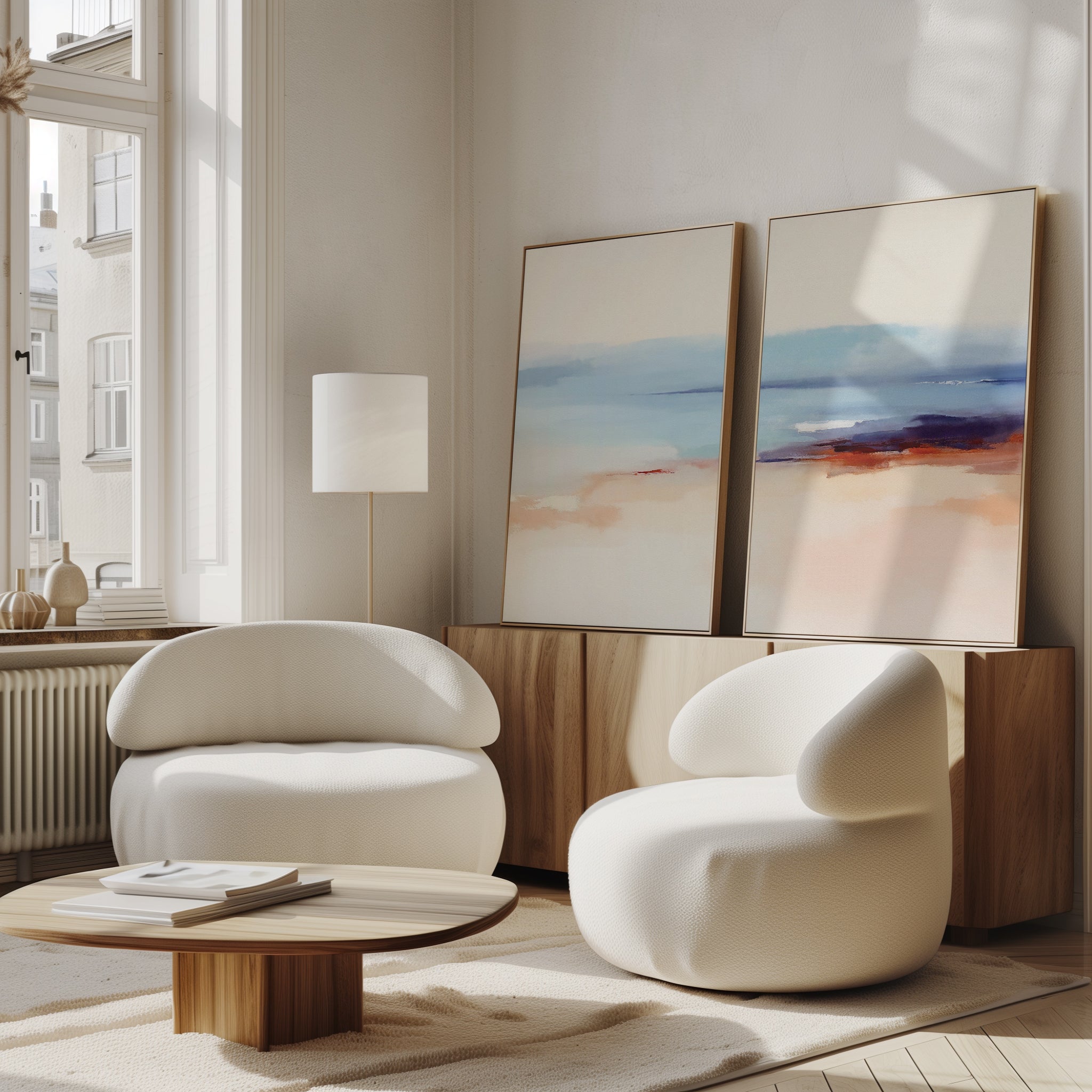 A modern living room features two rounded white armchairs, a low wooden coffee table, a floor lamp, and the Windswept Coastline - Set Of 2 wall art set atop a wooden sideboard against a white wall, brightened by large windows.