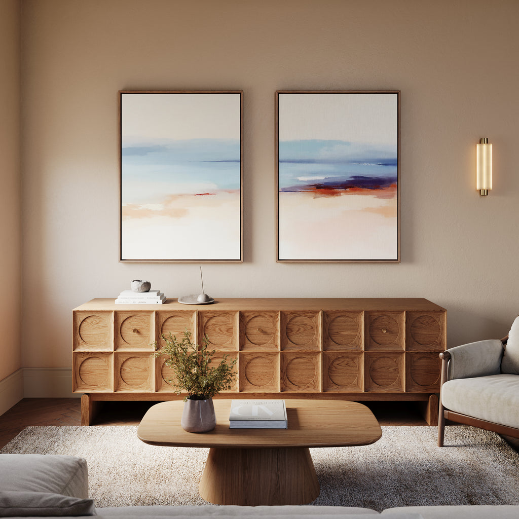 A modern living room features a wooden coffee table and sideboard, Windswept Coastline - Set Of 2 coastal wall art above the sideboard, a plant in a vase, neutral walls, and a cozy armchair.