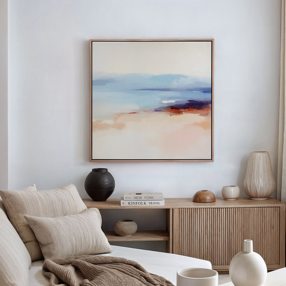 A modern living room with a beige sofa, neutral pillows, and wooden sideboard features minimalist decor—vases, books, and the Windswept Coastline Canvas Art - Square in soft pastels displayed on a white wall.