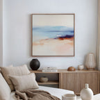 A modern living room with a beige sofa, neutral pillows, and wooden sideboard features minimalist decor—vases, books, and the Windswept Coastline Canvas Art - Square in soft pastels displayed on a white wall.