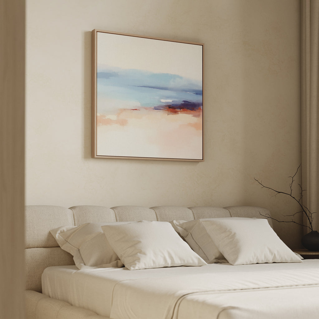 A minimalist bedroom with a beige upholstered bed, white pillows, and a soft white blanket. Above the bed hangs the Windswept Coastline Canvas Art - Square in pastel blue, pink, and orange. A bare branch decorates the side table.