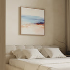 A minimalist bedroom with a beige upholstered bed, white pillows, and a soft white blanket. Above the bed hangs the Windswept Coastline Canvas Art - Square in pastel blue, pink, and orange. A bare branch decorates the side table.